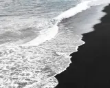 Kobieta w białej koszuli idzie po czarnych plażach, zostawiając ślady na piasku. Fale oceanu delikatnie obmywają brzeg.