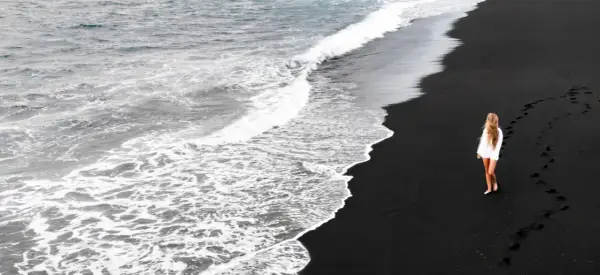 Kobieta w białej koszuli idzie po czarnych plażach, zostawiając ślady na piasku. Fale oceanu delikatnie obmywają brzeg.